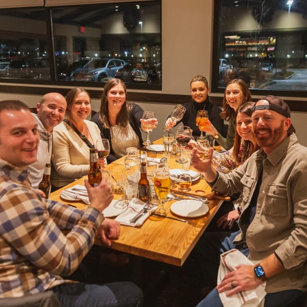 A group of eight smiling friends toasting with drinks at a restaurant table at night.