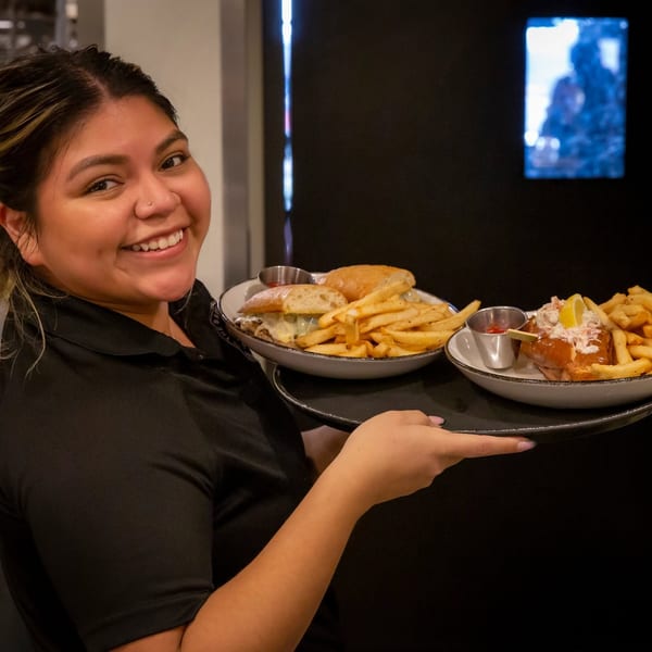 Smiling restaurant server holding a tray with two plates of sandwiches and fries.
