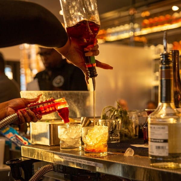 Hands pouring dark and red liquids into ice-filled glasses at a warm, inviting bar.